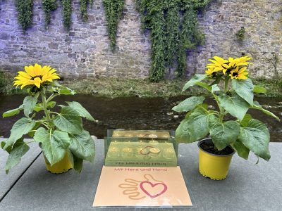 Auf der Erftmauer stehen zwei Sonnenblumen; in dazwischen eine Reihe von Teelichtern und eine Tafel mit der Inschrift "Mit Herz und Hand"