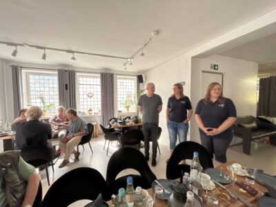 Drei Personen stehen, andere sitzen im Hintergrund am Tisch.